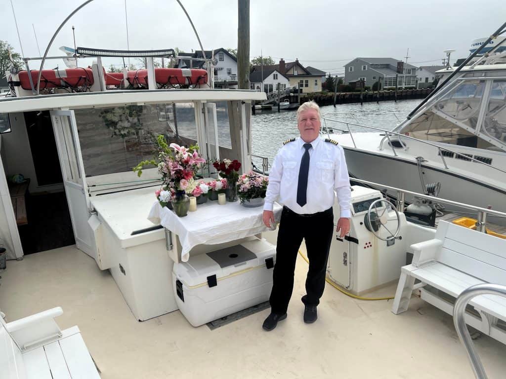 A man in a white shirt, black tie, and black pants stands on a boat. Behind him are a table with floral arrangements and various boats docked near a waterfront neighborhood in Queens County, known for its serene pet sea burials. The sky is overcast.