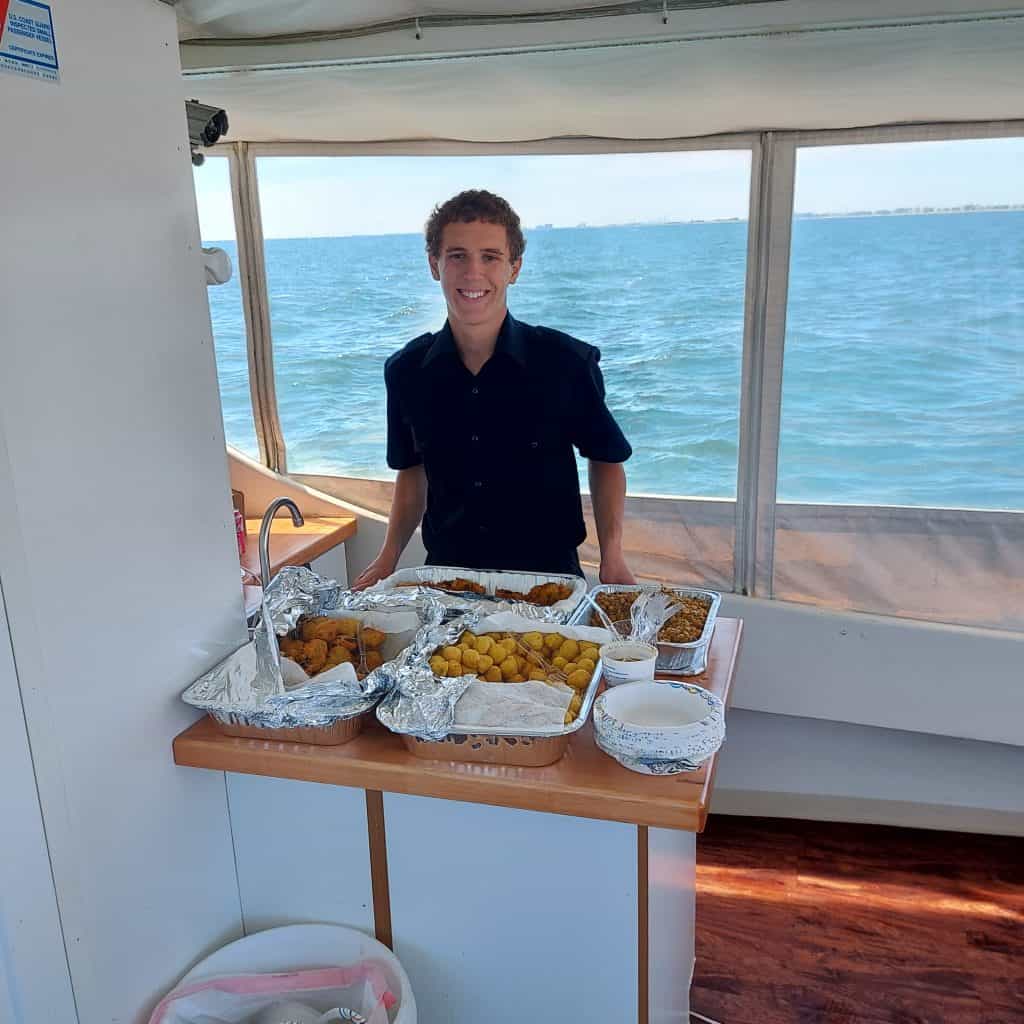 A smiling person sits behind a table of food on a boat at sea, ideal for Sea Burials Long Island.