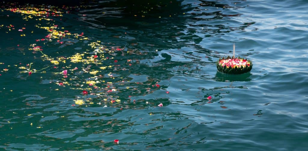 A circular krathong adorned with flowers floats on the water's surface, surrounded by scattered colorful petals. The water shimmers with light reflections, creating a serene and festive atmosphere reminiscent of an unattended sea burial in Nassau County.