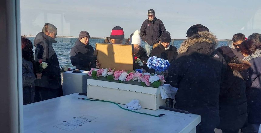 A group gathers for a sea burial off Long Island on a boat with a flower-adorned casket.