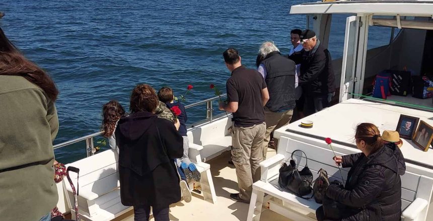 A group of people gathered on a boat off the coast for an unattended sea burial in Nassau County. Some are sitting on benches while others stand, looking out over the water. A few individuals hold or throw flowers into the ocean. One person's finger partially covers the top right corner of the image.